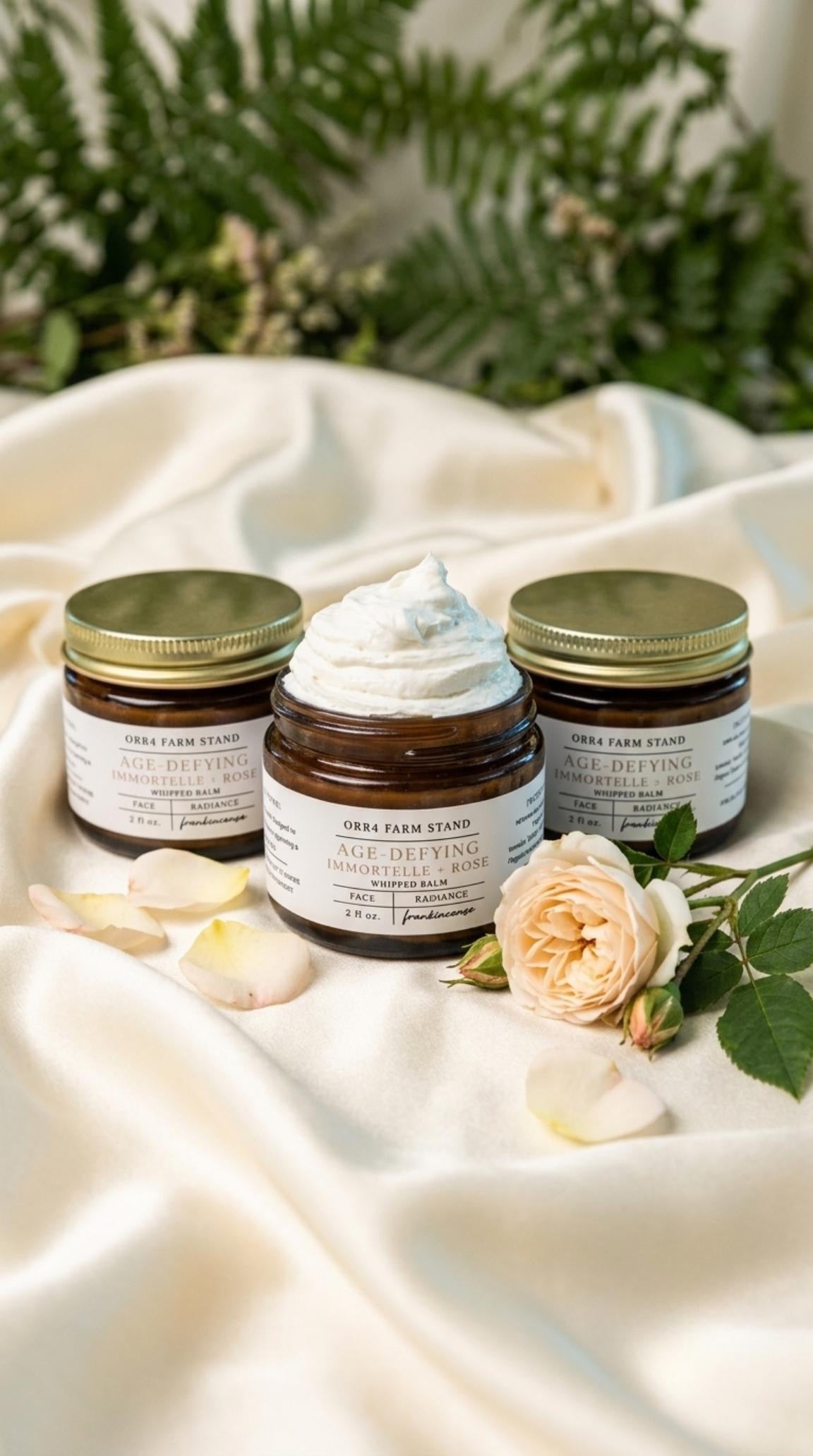 Immortelle and Rose facial balm jars arranged on cream fabric with roses and greenery.