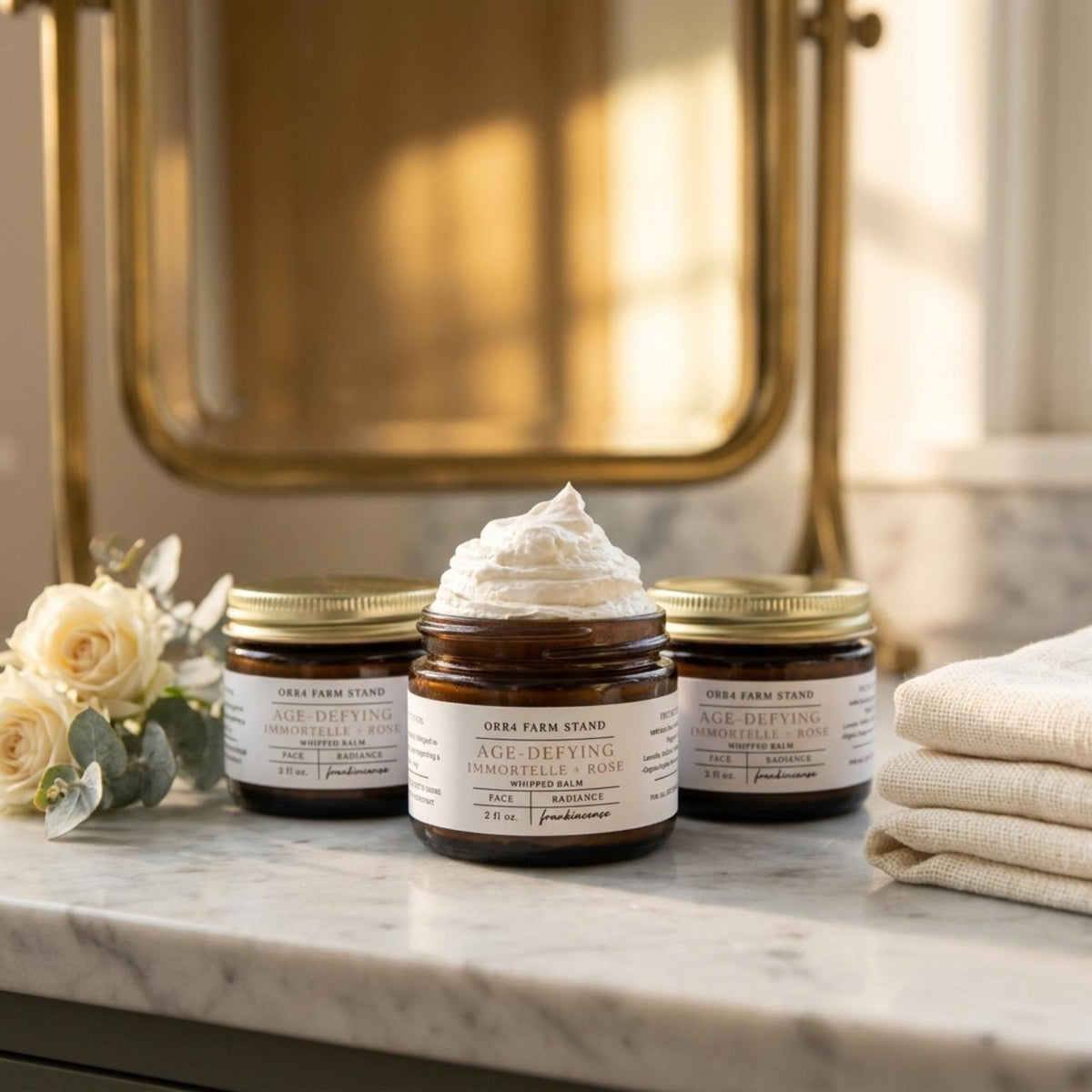Immortelle and Rose facial balm jars on a bathroom counter with roses, towels and an open jar of balm.