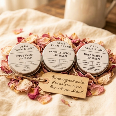 Three Orr4 Farm Stand lip balm tins on linen among dried rose petals, with a tag reading Pure ingredients, luxurious care.